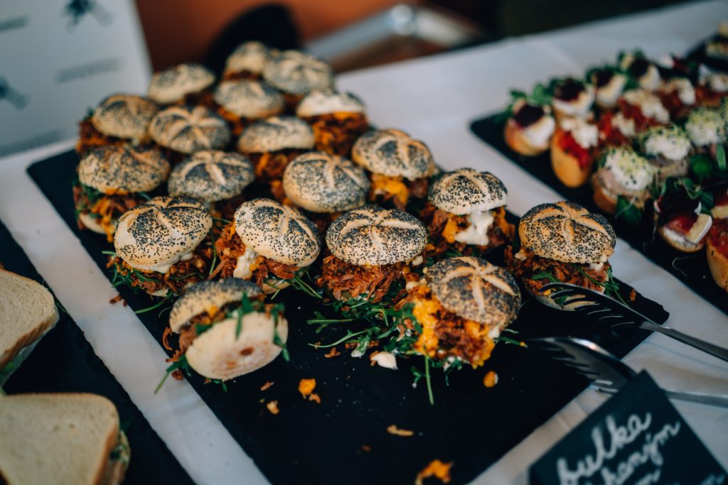 Mini burger bulky sypané mákem, plněné trhaným masem, sýrem a rukolou, naaranžované na černé podložce na cateringové akci.